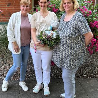 Drei Frauen posieren lächelnd vor einem Backsteingebäude, während die mittlere stolz einen Blumenstrauß hält.