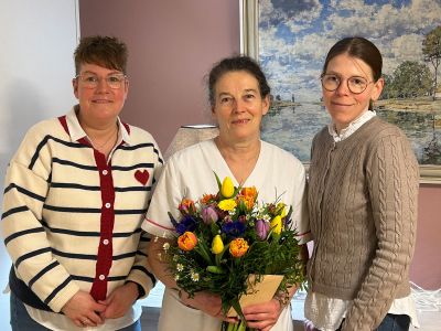 Drei Frauen stehen lächelnd zusammen, während eine von ihnen einen bunten Blumenstrauß in den Händen hält.