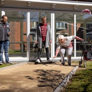 Drei Personen genießen einen sonnigen Tag im Freien, während eine ältere Frau mit einem Rollator spielt. | © Name