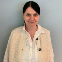 Eine Frau in weißem Hemd und Strickweste lächelt vor einer hellblauen Wand.