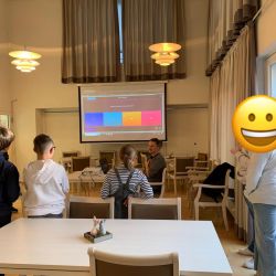 In einem hellen Raum beobachten mehrere Kinder einen Bildschirm, während ein lächelnder Erwachsener vorne sitzt.