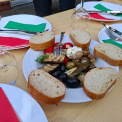 Ein Teller mit Antipasti, Brot und Oliven steht auf einem Tisch mit bunten Servietten und umgedrehten Weingläsern.