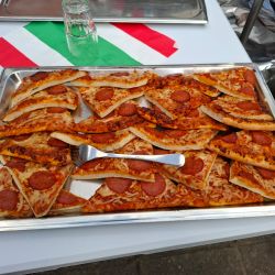 Auf einem Tisch liegen viele Salami-Pizzastücke auf einem Blech, begleitet von einer italienischen Flagge im Hintergrund.