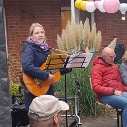 Eine Frau spielt Gitarre und singt in einer gemütlichen Outdoor-Veranstaltung mit bunten Laternen im Hintergrund.