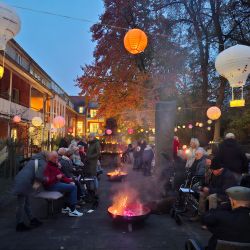 Menschen versammeln sich bei Dämmerung um Feuerstellen, umgeben von Laternen, in einem herbstlichen Garten.