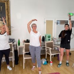 Drei Frauen heben ihre Arme in einem hellen Raum, während sie eine Yogaübung gemeinsam ausführen.