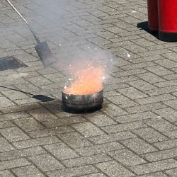 Ein Topf mit glühendem Metall sprüht Funken auf den Pflasterboden, während ein Werkzeug daneben liegt.