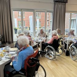 Senioren in Rollstühlen unterhalten sich beim Essen in einem hellen Raum mit großen Fenstern.