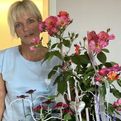 Eine Frau im blauen T-Shirt steht neben einem üppigen Blumenarrangement mit rosa Blüten und einem silbernen Herzanhänger.
