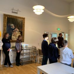 Eine Gruppe von Menschen betrachtet aufmerksam ein großes Gemälde im Raum, während ein Servierwagen daneben steht.