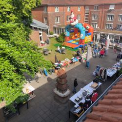 Ein bunter Flohmarkt mit Hüpfburg und Ständen voller Kleidung und Spielzeug belebt einen sonnigen Innenhof.