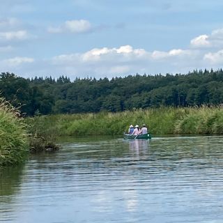Drei Personen paddeln in einem Kanu auf einem ruhigen Fluss, umgeben von üppigem Grün und blauem Himmel.