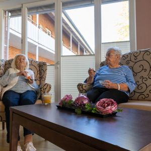 Zwei ältere Frauen genießen Snacks und Unterhaltung in einem gemütlichen Raum mit Pflanzen und einem Tisch.
