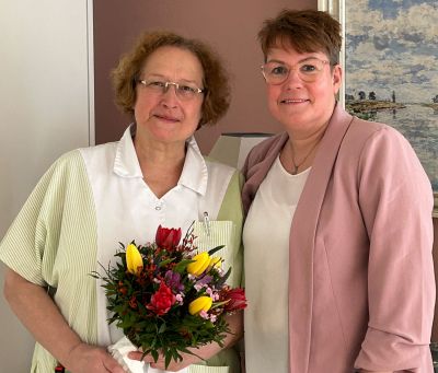 Zwei Frauen, eine mit einem Blumenstrauß, posieren freundlich in einem hellen Raum für ein gemeinsames Foto.