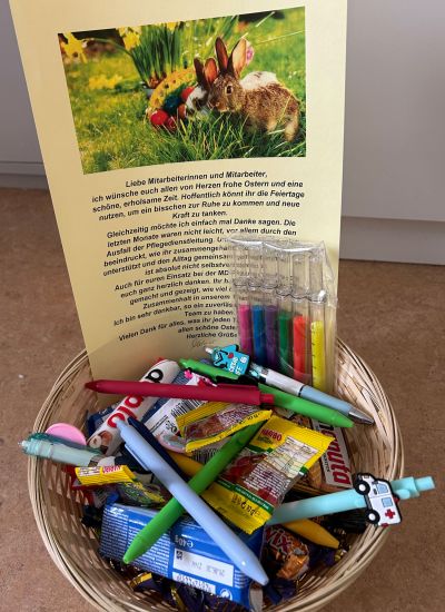 Ein Korb voller Süßigkeiten und bunter Stifte steht neben einem Ostergruß mit zwei Hasen auf einer Wiese.