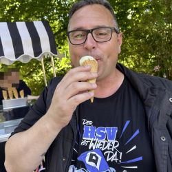 Ein Mann genießt ein Eis vor einem Eiswagen im Sommer und trägt stolz ein HSV-Fan-T-Shirt.