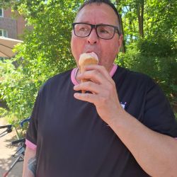 Ein Mann genießt ein Eis im Freien, umgeben von grünen Bäumen und sonnigem Wetter. | © Name
