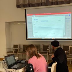 Zwei Personen arbeiten an Laptops in einem Seminarraum mit Projektion auf der Leinwand.