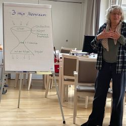 Eine Frau erklärt vor einem Flipchart mit der Überschrift "3 min Atemraum" in einem hellen Raum.