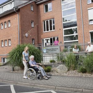 Eine Frau schiebt eine Seniorin im Rollstuhl vor dem Posener Altenheim, während andere Bewohner draußen entspannen.