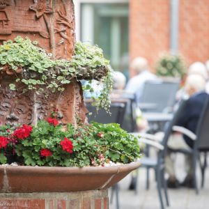 Blühende Geranien und eine kunstvolle Steinskulptur zieren den Garten, während Menschen entspannt im Hintergrund sitzen.