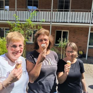 Drei Frauen genießen bei sonnigem Wetter ein Eis und lächeln fröhlich vor einem Backsteingebäude in die Kamera.