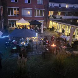 Im Innenhof eines Gebäudes sitzen Menschen gemütlich um ein Lagerfeuer, während bunte Lichter die Szene erhellen.