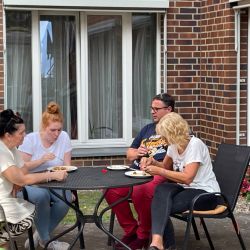 Vier Personen genießen ein gemeinsames Essen an einem runden Tisch vor einem schönen Backsteinhaus im Freien.