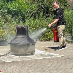 Ein Mann löscht im Freien mit einem Feuerlöscher ein Feuer neben einem kleinen Metallofen auf einer gepflasterten Fläche.