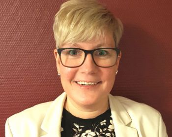 Eine lächelnde Frau mit Brille und kurzem blondem Haar trägt einen cremefarbenen Blazer vor einer roten Wand.