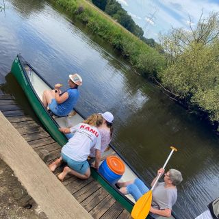 Drei Personen steigen am Flussufer in ein Kanu ein, bereit für eine entspannte Kanutour an einem ruhigen Sommertag.