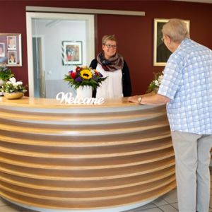 Ein älterer Mann spricht mit einer Frau an einem modernen Empfangstresen, dekoriert mit Blumen und einem "Welcome"-Schild.