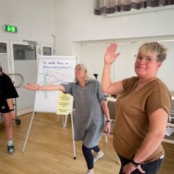 Drei Frauen machen entspannende Übungen vor einem Flipchart, um Stressbewältigung im Workshop zu lernen.