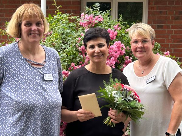 Drei Frauen posieren lächelnd vor blühenden Pflanzen, eine hält einen Blumenstrauß und einen Umschlag. | © Name
