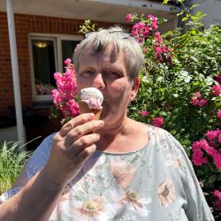 Eine Frau genießt ein Eis in der Sonne, umgeben von blühenden pinken Blumen im Garten.