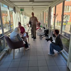 Drei Personen in einem hellen Flur unterhalten sich, während ein Kamerateam Aufnahmen macht. | © Name