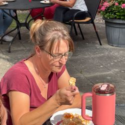 Eine Frau in einem roten Shirt genießt draußen ein Stück Kuchen neben einer rosa Thermoskanne auf der Terrasse.