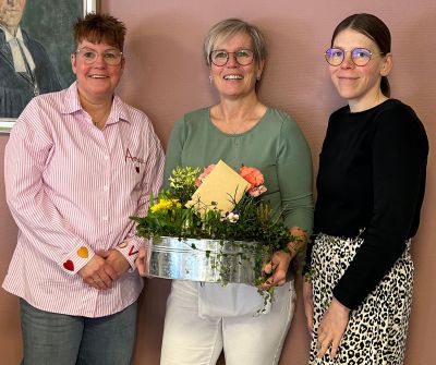Drei Frauen lächeln fröhlich, während sie einen blumigen Geschenkkorb für einen besonderen Anlass präsentieren.