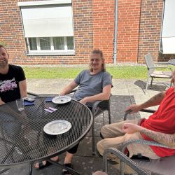 Drei Männer sitzen entspannt an einem runden Tisch vor einem Backsteingebäude und genießen den sonnigen Tag.