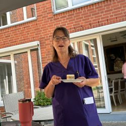 Eine Frau in lila Uniform hält ein Stück Kuchen auf einem Teller vor einem roten Backsteingebäude.