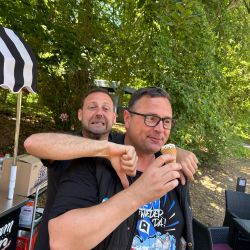 Zwei Männer stehen im Grünen, einer genießt ein Eis lächelnd, während der andere mit einem Daumen nach unten scherzt.