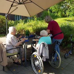Unter einem Sonnenschirm genießen zwei ältere Frauen ein Essen im Freien, während eine Betreuerin ihnen hilft.