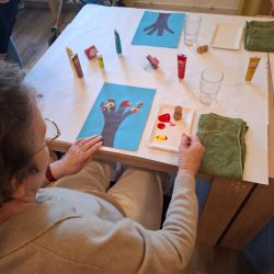 Eine ältere Dame malt mit bunten Farben auf Papier, um einen kreativen Baum zu gestalten.