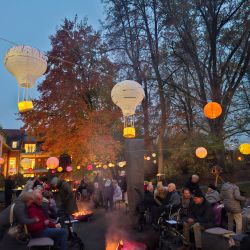 Ein gemütliches Herbstfest am Abend mit leuchtenden Laternen und Menschen, die um Feuerstellen versammelt sind.