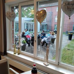 Durch das Fenster plaudern Menschen entspannt im Garten eines Backsteingebäudes bei Getränken und genießen die Atmosphäre.