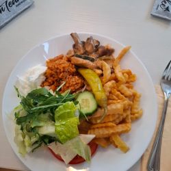 Ein Teller mit Pommes, Salat, Reis, Pilzen und einer Peperoni liegt auf einem Tisch neben Besteck und Servietten.