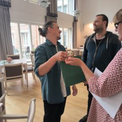 Drei Personen unterhalten sich fröhlich in einem Raum mit großen Fenstern und Holzfußboden.