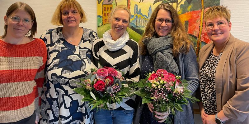 Fünf Frauen lächeln vor einem bunten Gemälde, während zwei Blumensträuße in den Händen halten.
