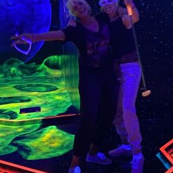 Zwei Personen in leuchtender Kleidung genießen Minigolf in einer neonbeleuchteten, galaktischen Umgebung voller Farben.