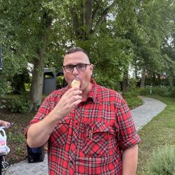 Ein Mann in einem roten Karohemd genießt ein Eis unter den grünen Bäumen im Freien.